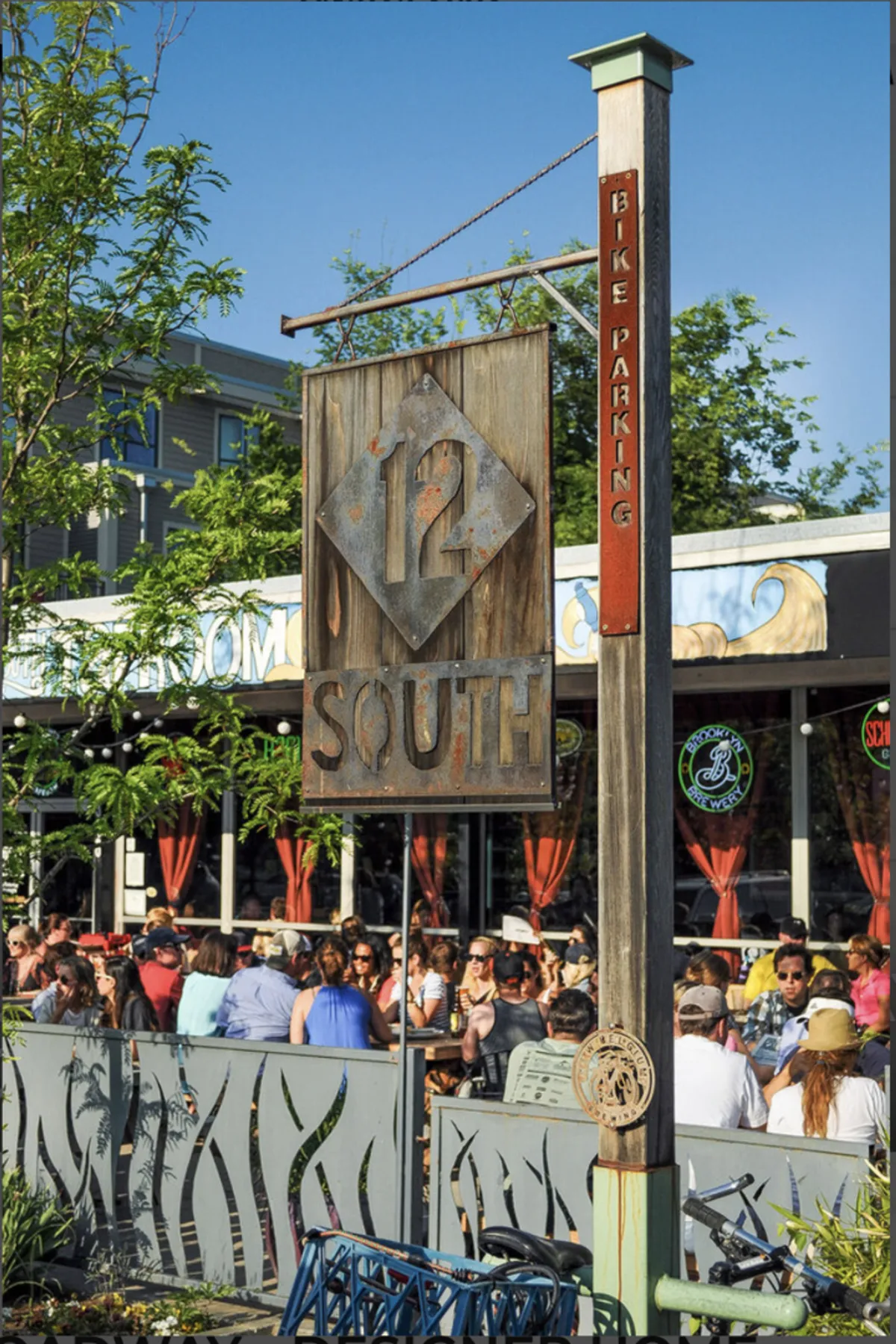A street-level exterior shot of the 12 South district, featuring the vibrant 12 South sign, a crowd of people dining outdoors, and a bike parking sign. The environment is lively, community-focused, and full of local flavor.