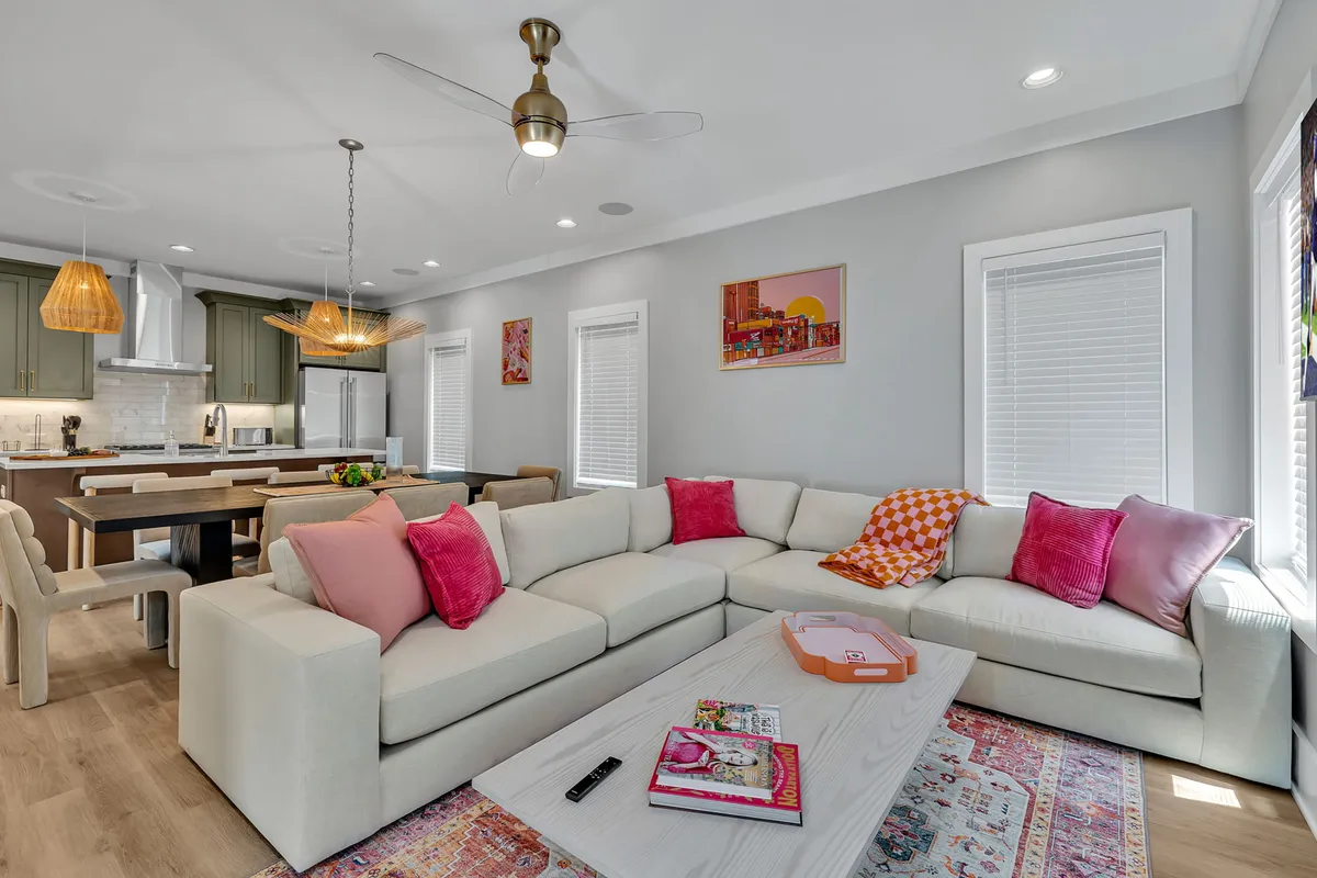 The image showcases a spacious, sunlit interior featuring a plush sofa adorned with colorful cushions, a large wooden dining table perfect for group meals, a modern kitchen island with bar stools, and vibrant wall art—all creating a chic, inviting atmosphere ideal for gatherings.