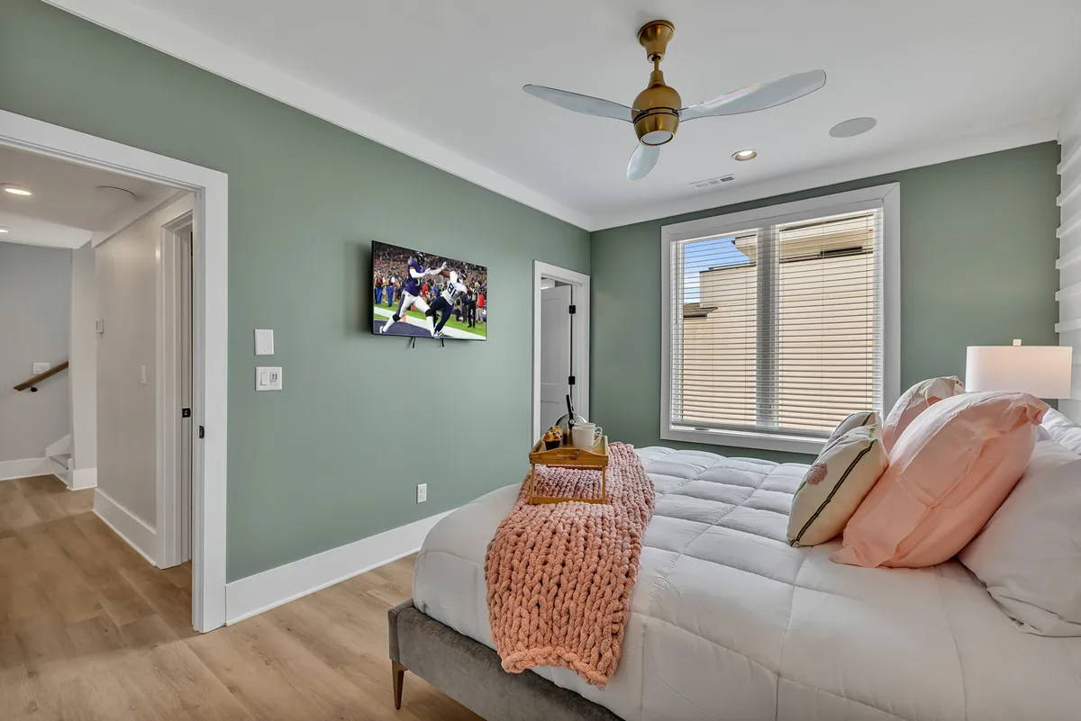 A cozy, modern bedroom featuring a bed with decorative pillows and a knitted throw blanket, a television mounted on the wall, and a large window with blinds, allowing in ample natural light.
