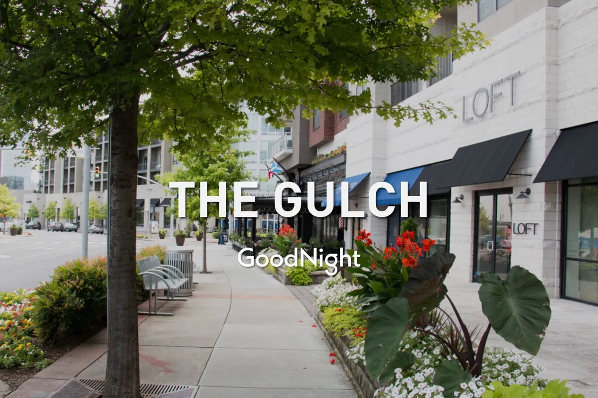 Storefronts line a lively street, bordered by flower beds, trees, and benches. The atmosphere is vibrant, suggesting a walkable shopping area ideal for people of all ages to explore, shop, and socialize.
