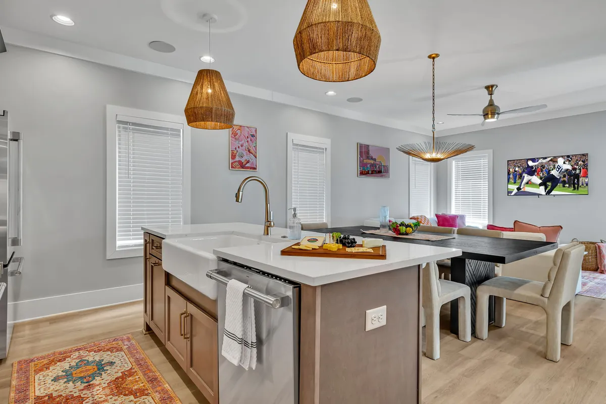A wide-angle view of an open-concept main living area featuring a modern kitchen with a prominent island and pendant lights, a spacious dining area set for eight, colorful artwork, and a comfortable lounge space with a large television. The seamless flow between kitchen, dining, and living zones is clearly visible.
