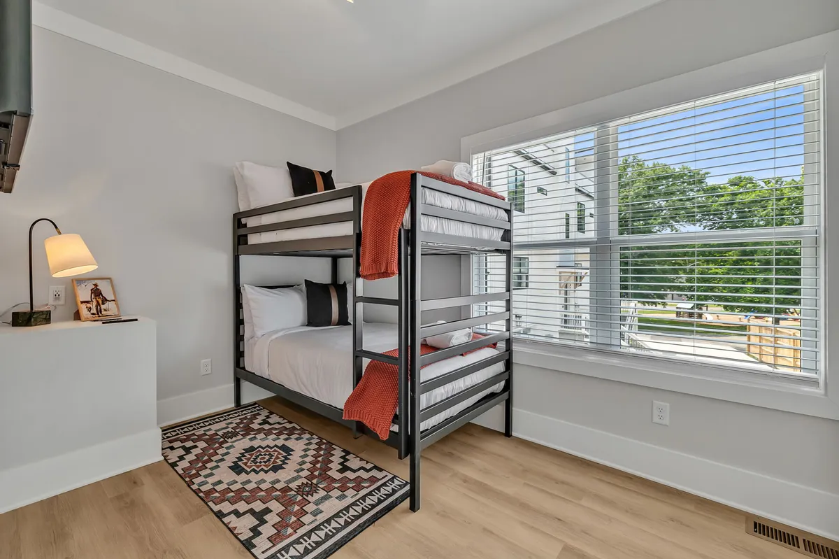 A cozy bunk room with a sturdy bunk bed, a soft rug, a lamp, and a window that lets in natural light. The space feels playful and perfect for children or group sleepovers.
