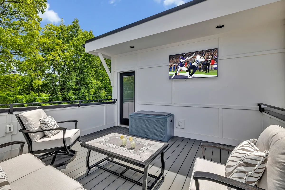 A stylish rooftop patio featuring a cushioned seating area, an outdoor television mounted on the wall, a decorative table, and greenery in the background. The setting is relaxed and designed for both entertaining and personal enjoyment.