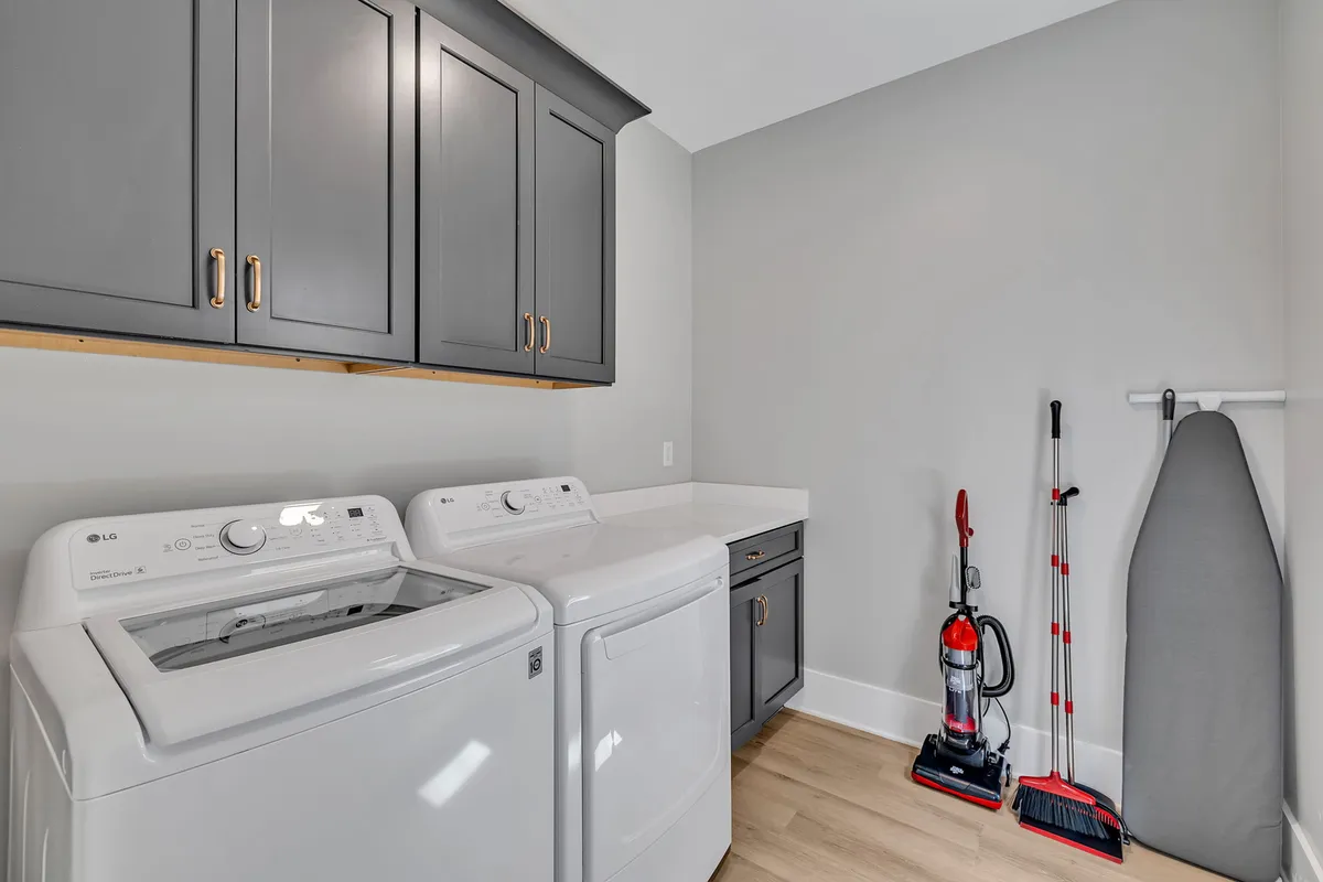 A functional laundry room with a washer and dryer set beneath upper cabinets, a vacuum cleaner, and an ironing board, creating an orderly and well-equipped cleaning zone.