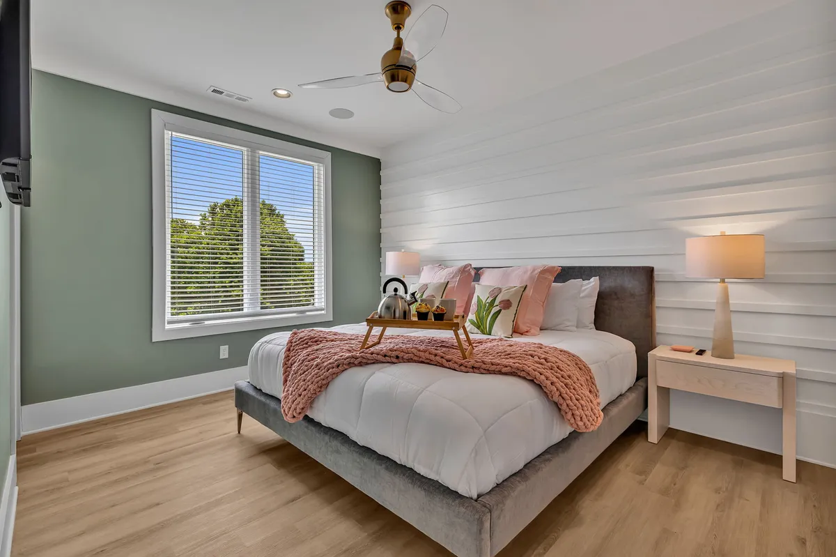 A bright, welcoming bedroom with a bed adorned with a pink throw blanket and decorative pillows, a nightstand with a lamp, and windows that bathe the room in natural light.