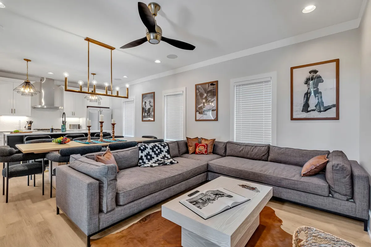 A cozy, stylish living and dining area featuring a large sectional sofa perfect for group lounging, a modern chandelier, a dining table set for eight, and vibrant artwork on the walls. The arrangement encourages both casual socializing and formal gatherings.